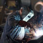 Welder using a MIG welder to join metal in a workshop, with sparks visible around the welding area.